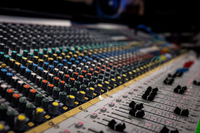 Close-up van een mengtafel vol gekleurde knoppen en faders, podium wazig op de achtergrond